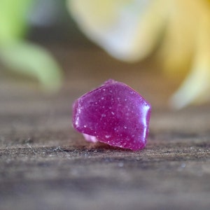 Op de afbeelding: Een diep roze, gefacetteerde edelsteen met een ruw, getextureerd oppervlak. De edelsteen ligt op een houten oppervlak.