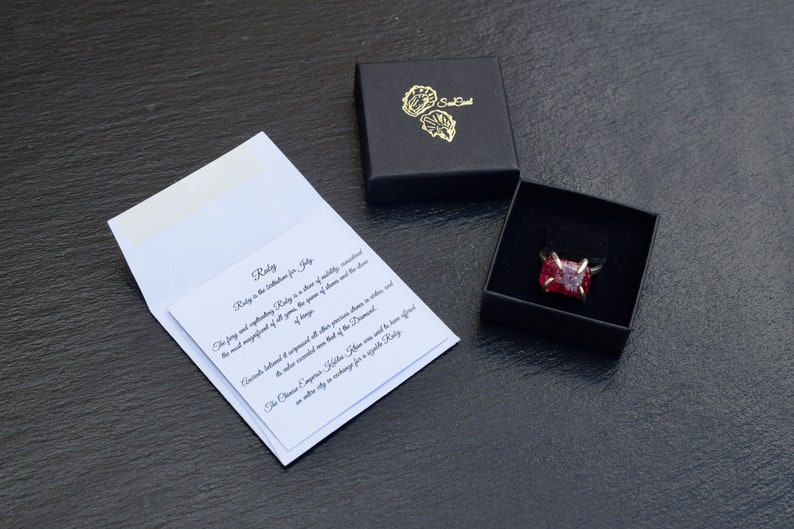 Op de afbeelding: Een ring met een rode edelsteen in een zwarte fluwelen doos met een wit kaartje waarop staat: "Robijn is de geboortesteen voor juli. De vurige en boeiende robijn is een steen van vitaliteit, passie en de meest magnifieke van alle edelstenen. De koningin van de edelstenen en de steen die als uitgebalanceerd en onovertroffen wordt beschouwd van alle andere edelstenen in kleur en glans. Men beschouwde de diamant als de enige steen die waardig was voor een geschenk voor een koning. De Chinese keizer Kublai Khan kreeg te horen dat hij een hele stad moest uitgeven in ruil voor &eacute;&eacute;n enkele robijn."