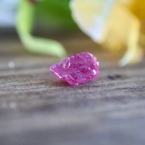 Op de afbeelding: Een kleine, ruwe, roze robijn edelsteen op een houten oppervlak.