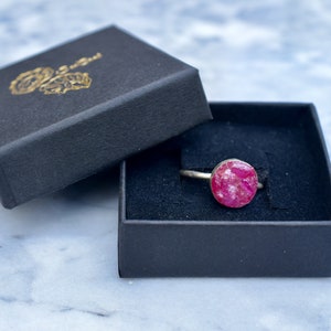 May include: A pink gemstone ring in a black ring box. The ring has a silver band and a round pink gemstone.