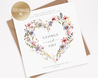 May include: A white card with a floral heart design. The heart is made of pink, purple, and blue flowers. The text "FOR A VERY SPECIAL SON AND DAUGHTER IN LAW" is at the top of the card. The names "GEORGE and AMY" are in the center of the heart. The text "ON YOUR WEDDING DAY 13.07.24" is at the bottom of the card.