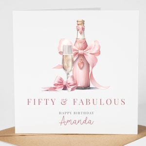 May include: A pink birthday card with a bottle of champagne and a glass of champagne. The bottle is tied with a pink ribbon and a heart charm. The card says "Fifty & Fabulous" and "Happy Birthday Amanda".