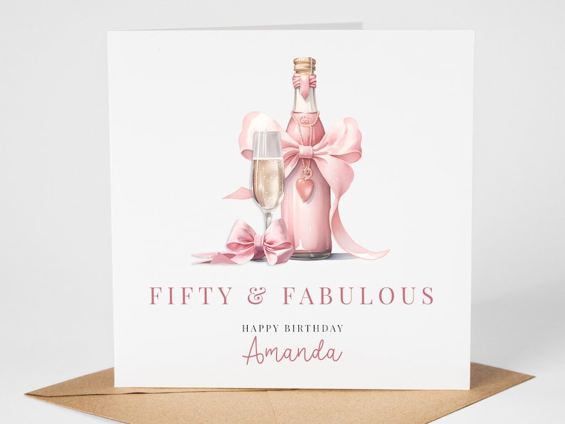 May include: A pink birthday card with a bottle of champagne and a glass of champagne. The bottle is tied with a pink ribbon and a heart charm. The card says "Fifty & Fabulous" and "Happy Birthday Amanda".