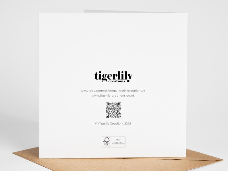 May include: A white greeting card with the text "tigerlily creations" in black. Below the text are website addresses and a QR code. The card is on a brown envelope.