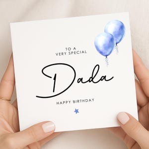 May include: A white square birthday card with the words "TO A VERY SPECIAL Dada HAPPY BIRTHDAY" in black script. Two blue watercolour balloons are in the upper right corner, and a blue star is at the bottom. The card is held by hands.