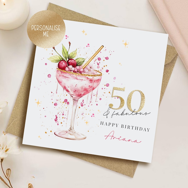 May include: A birthday card with a watercolor illustration of a pink cocktail with cherries and a gold rim. The card reads "50 & fabulous, Happy Birthday Ariana" in gold and pink script. A gold foil sticker says "Personalise Me".