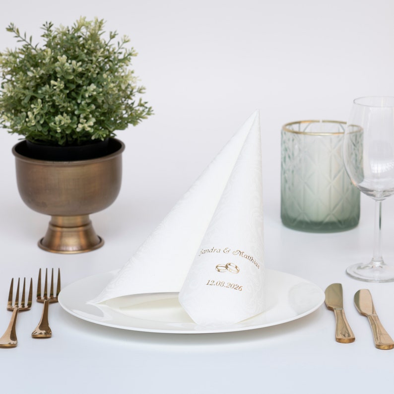 Puede incluir: Servilleta blanca doblada con letras doradas que dicen "Sandra & Matthew 12.08.2026" y dos anillos dorados. La servilleta est&aacute; en un plato blanco con cubiertos dorados.