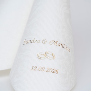 Puede incluir: Servilleta de boda blanca con letras doradas. La servilleta dice "Sandra & Matthias" y "12.08.2026" con dos anillos dorados entre los nombres y la fecha.