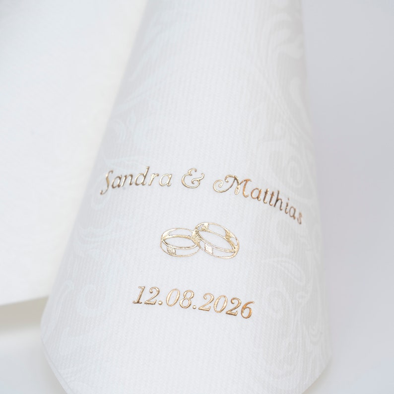 Puede incluir: Servilleta de boda blanca con letras doradas. La servilleta dice "Sandra & Matthias" y "12.08.2026" con dos anillos dorados entre los nombres y la fecha.