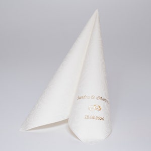 Puede incluir: Servilleta de boda blanca con un patr&oacute;n en relieve. La servilleta est&aacute; doblada en forma de tri&aacute;ngulo. El texto "Sandra & Matthew" y "12.08.2026" est&aacute; impreso en dorado en la servilleta, junto con un dise&ntilde;o de anillo dorado.