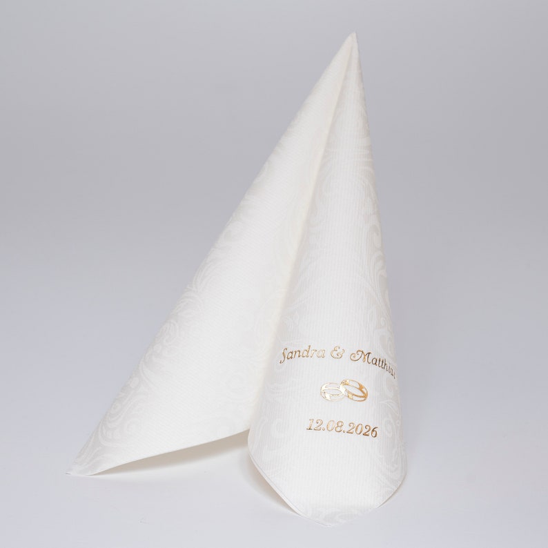 Puede incluir: Servilleta de boda blanca con un patr&oacute;n en relieve. La servilleta est&aacute; doblada en forma de tri&aacute;ngulo. El texto "Sandra & Matthew" y "12.08.2026" est&aacute; impreso en dorado en la servilleta, junto con un dise&ntilde;o de anillo dorado.