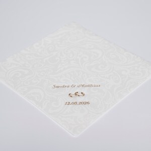 Puede incluir: Invitaci&oacute;n de boda blanca con un patr&oacute;n de damasco blanco. La invitaci&oacute;n dice "Sandra & Matthias" con dos anillos dorados y la fecha "12.08.2026".