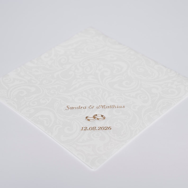 Puede incluir: Invitaci&oacute;n de boda blanca con un patr&oacute;n de damasco blanco. La invitaci&oacute;n dice "Sandra & Matthias" con dos anillos dorados y la fecha "12.08.2026".