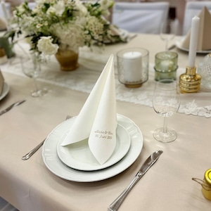 Puede incluir: Una servilleta blanca doblada con el texto "Alicia & Steve 17. Juni 2023" en un plato blanco con borde dorado. El plato est&aacute; sobre un mantel beige con una sobreposici&oacute;n de encaje blanco. Hay un vaso de agua y un cuchillo de plata en la mesa.