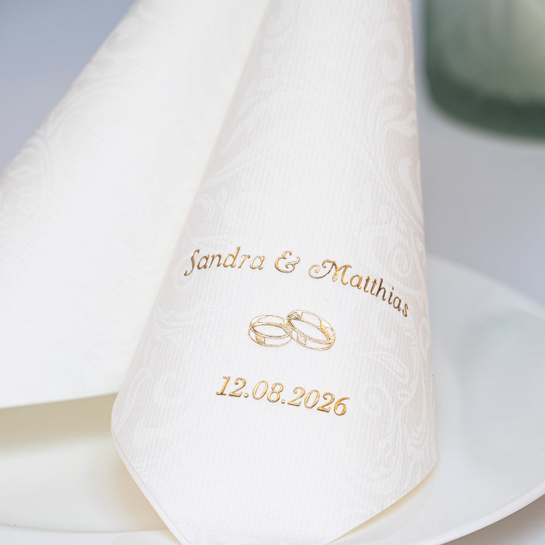 Puede incluir: Servilleta de boda blanca con letras doradas que dicen "Sandra & Matthias" y "12.08.2026". La servilleta tambi&eacute;n presenta una ilustraci&oacute;n dorada de dos anillos entrelazados.