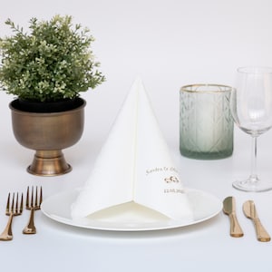 Puede incluir: Una servilleta blanca doblada con el texto "Sandra & Thomas" y la fecha "12.08.2021" impresa en dorado. La servilleta est&aacute; sobre un plato blanco con cubiertos dorados y un portavelas de vidrio verde.
