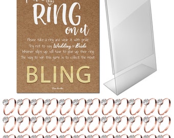 May include: A wedding game set with a brown card that reads "Put a Ring on It" and "Bling". Includes a clear acrylic sign holder and numerous rose gold rings with clear, round center stones.
