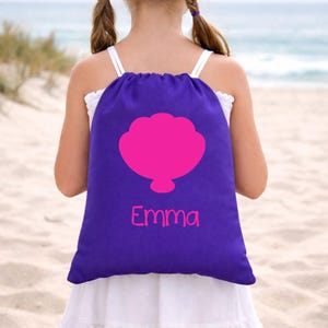 Puede incluir: Mochila morada con cordón, con un diseño de concha rosa y el nombre "Emma" en rosa. La bolsa se lleva sobre los hombros. El fondo es una escena de playa con arena y el mar.