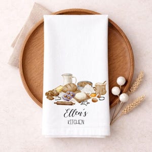 May include: White kitchen towel featuring a watercolour illustration of baking ingredients and tools, such as bread, flour, and a rolling pin. The text "Ellen's Kitchen" is printed at the bottom. The towel rests on a wooden tray.