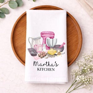 May include: White kitchen towel featuring watercolour illustrations of baking equipment, such as a pink stand mixer, a mortar and pestle, and a measuring jug. The text "Martha's Kitchen" is printed in black script.