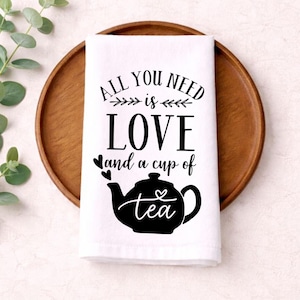 May include: White kitchen towel with the text "ALL YOU NEED is LOVE and a cup of tea" in black. The tea cup is a black silhouette. The towel is on a wooden tray.