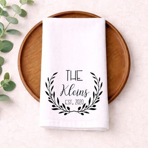 May include: White hand towel with a black floral wreath design and the text "THE Kleins EST. 2020". The towel is folded and placed on a wooden plate, with green leaves in the background.