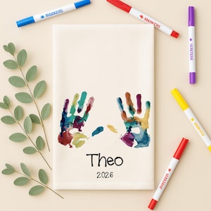 May include: A rectangular off-white fabric towel featuring two colorful handprints in shades of blue, green, red, and yellow. Below the handprints, the name Theo and the year 2026 are printed in black text. Five markers in blue, red, purple, yellow, and red surround the towel.