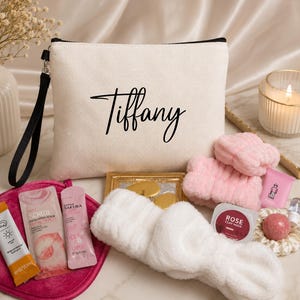 May include: A cream-coloured cosmetic bag with the name "Tiffany" in black script. The bag is surrounded by beauty products, including a pink headband, face scrub, and a rose clay mask. A lit candle and flowers are in the background.