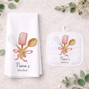 May include: White kitchen towel and pot holder set featuring a watercolour design of a whisk, spatula, and wooden spoon tied with a pink ribbon. The words "Nana's Kitchen" are written below the design.