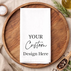 May include: A rectangular white kitchen towel featuring the black printed text YOUR Custom Design Here centred on the fabric. The towel is folded neatly and presented as a blank canvas for personalisation, showcasing a clean and simple aesthetic.