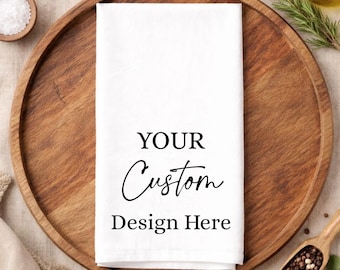 May include: A rectangular white kitchen towel featuring the black printed text YOUR Custom Design Here centered on the fabric. The towel is folded neatly and presented as a blank canvas for personalization, showcasing a clean and simple aesthetic.
