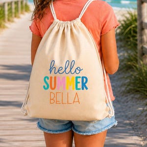 Może przedstawiać: Beżowy plecak ze sznurkiem z napisem hello SUMMER BELLA w niebieskich, pomarańczowych, żółtych i turkusowych literach. Torba ma białe sznurkowe paski i jest noszona na koralowym t-shircie oraz niebieskich szortach dżinsowych na tle plaży.