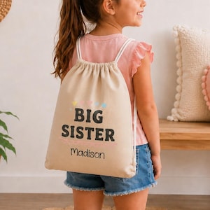 May include: A beige drawstring backpack with the words "BIG SISTER" in black, and the name "Madison" below. The text is accented with pastel hearts and stars. The backpack has white straps and is worn by a young girl.