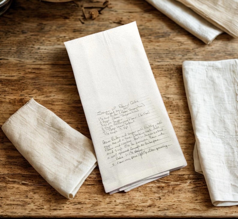 May include: A rectangular white kitchen towel featuring a handwritten recipe for Seven Up Pound Cake in black ink. The text includes ingredients like butter, sugar, eggs, flour, lemon extract, and 7 Up, along with baking instructions. The towel is folded.
