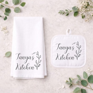 Puede incluir: Juego de paño de cocina y agarradera blancos. Ambos artículos presentan el texto "Tanya's Kitchen" en una fuente cursiva, enmarcado por un diseño botánico negro. La agarradera es cuadrada y tiene un lazo para colgar.