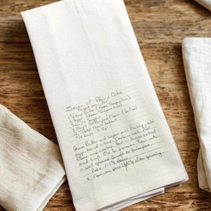 May include: A rectangular off-white kitchen towel featuring a printed handwritten recipe for Seven Up Pound Cake. The text includes ingredients like butter, sugar, flour, lemon extract, and 7-Up, along with baking instructions for a bundt or tube pan.