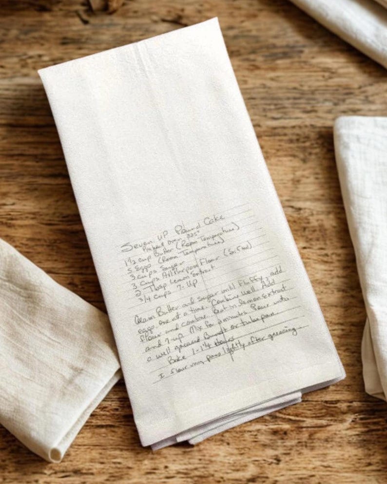 May include: A rectangular off-white kitchen towel featuring a printed handwritten recipe for Seven Up Pound Cake. The text includes ingredients like butter, sugar, flour, lemon extract, and 7-Up, along with baking instructions for a bundt or tube pan.