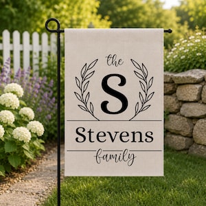 May include: A beige garden flag with black text and a black metal stand. The flag features a large letter "S" within a laurel wreath, the name "Stevens", and the word "family". The flag is displayed outdoors in a garden setting.