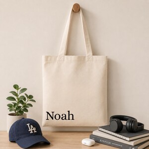 Pu&ograve; includere: Una borsa tote in tela beige con il nome Noah stampato in un carattere serif nero. La borsa ha due lunghi manici ed &egrave; mostrata appesa. Il design &egrave; semplice, funzionale e realizzato in un tessuto di colore chiaro con una forma rettangolare.