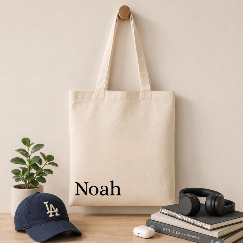 Pu&ograve; includere: Una borsa tote in tela beige con il nome Noah stampato in un carattere serif nero. La borsa ha due lunghi manici ed &egrave; mostrata appesa. Il design &egrave; semplice, funzionale e realizzato in un tessuto di colore chiaro con una forma rettangolare.