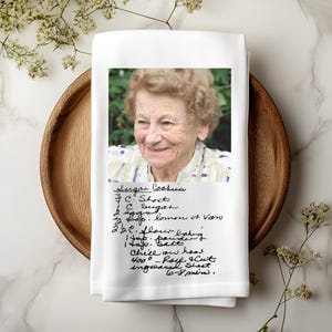 May include: White kitchen towel with a printed photo of an elderly woman and a handwritten sugar cookie recipe. The towel is on a wooden tray, with floral accents.
