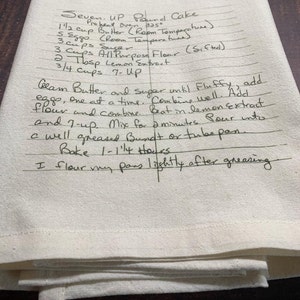 May include: A cream-coloured kitchen towel featuring a handwritten recipe for Seven-Up Pound Cake in black ink. The text includes ingredients like butter, eggs, sugar, flour, lemon extract, and 7-Up, along with baking instructions for a Bundt or tube pan.