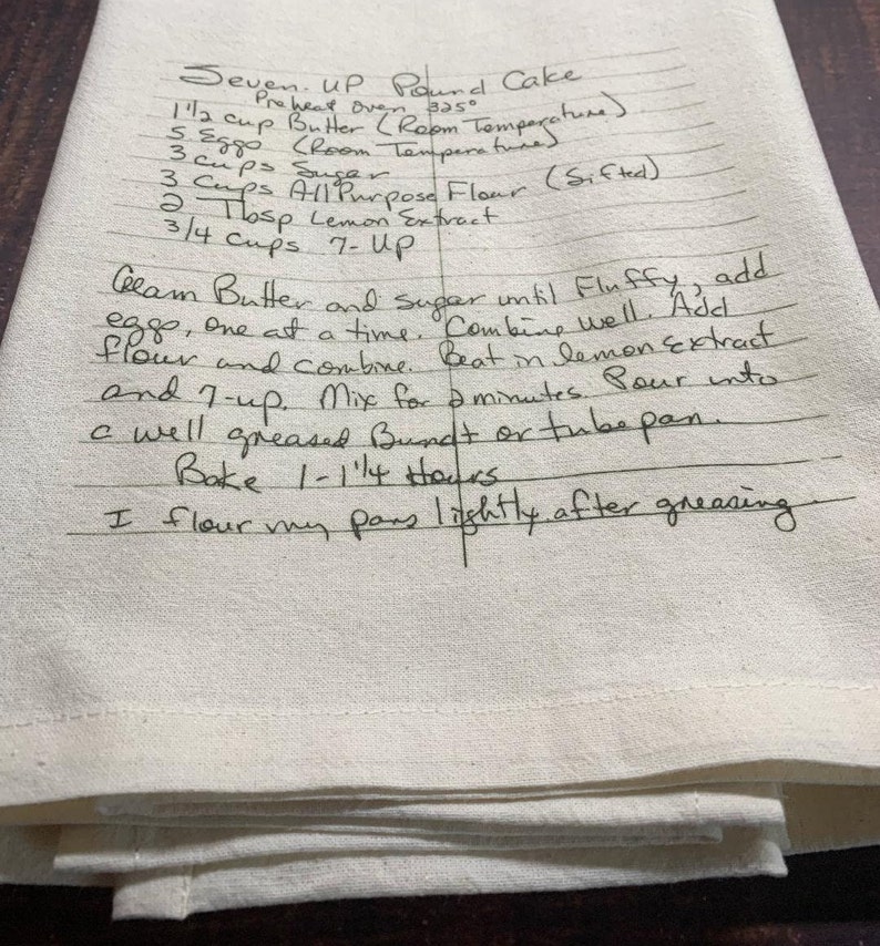 May include: A cream-coloured kitchen towel featuring a handwritten recipe for Seven-Up Pound Cake in black ink. The text includes ingredients like butter, eggs, sugar, flour, lemon extract, and 7-Up, along with baking instructions for a Bundt or tube pan.