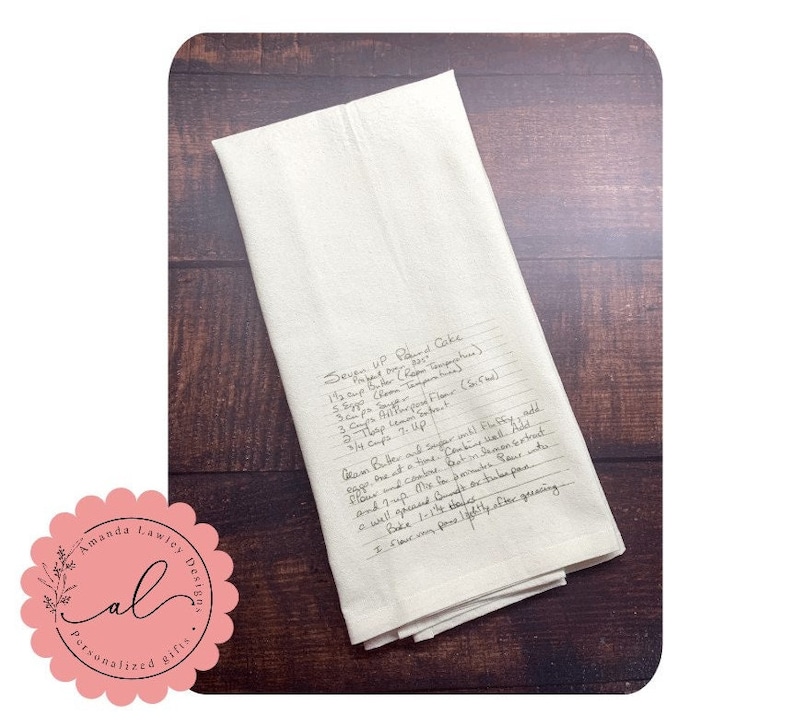May include: A folded off-white kitchen towel featuring a handwritten recipe for Seven Up Pound Cake in black ink. The text includes ingredients like 1 1/2 cups butter, 3 cups sugar, 3 cups all-purpose flour, 2 tbsp lemon extract, and 3/4 cups 7-Up.