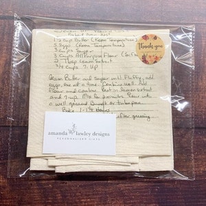 May include: A cream-coloured fabric tea towel featuring a handwritten recipe for Seven-Up Pound Cake in black ink. A circular sticker with floral graphics and the text Thank you is attached. A white card with the text amanda lawley designs is included.