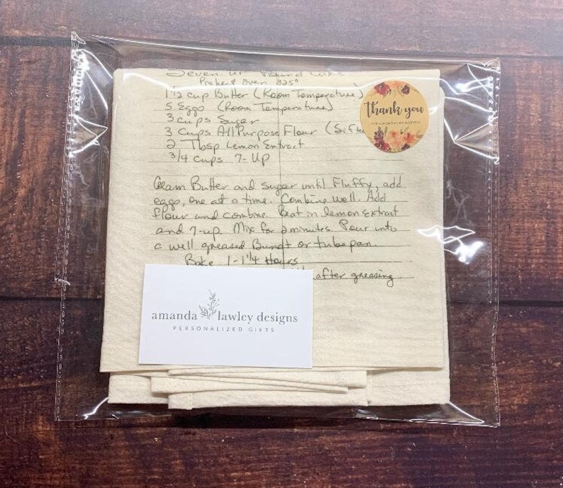 May include: A cream-coloured fabric tea towel featuring a handwritten recipe for Seven-Up Pound Cake in black ink. A circular sticker with floral graphics and the text Thank you is attached. A white card with the text amanda lawley designs is included.