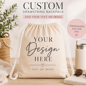 May include: A beige custom drawstring backpack with white drawstrings. The text "YOUR Design HERE" is printed in black script. Additional text includes "CUSTOM DRAWSTRING BACKPACK" and "ADD YOUR TEXT OR IMAGE". A pink circle reads "PERSONALIZED JUST FOR YOU!"
