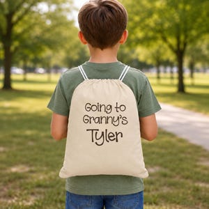 May include: A beige drawstring backpack with white straps is worn by a person. The backpack has the text "Going to Granny's" above the name "Tyler" in a black, handwritten-style font. The person is wearing a green shirt and blue jeans.
