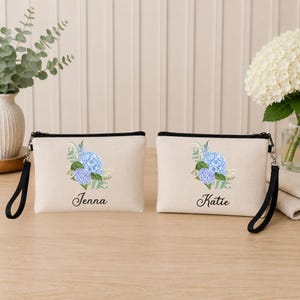 Puede incluir: Dos neceseres beige con cremallera, ribete negro y correas de muñeca. Cada neceser presenta un diseño de acuarela de hortensias azules y moradas con los nombres "Jenna" y "Katie" impresos debajo. Los neceseres están sobre una superficie de madera.