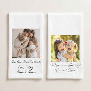 May include: Two white rectangular towels with printed family photos. One towel has a family photo with the text "We Love You So Much! Ben, Haley, Liam & Wren." The other towel has a photo of two girls with the text "We Love You, Grammy! Emma & Olivia."